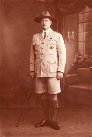 A man in a historical, military-style uniform with medals stands indoors holding a cane, wearing shorts and knee-high socks.
